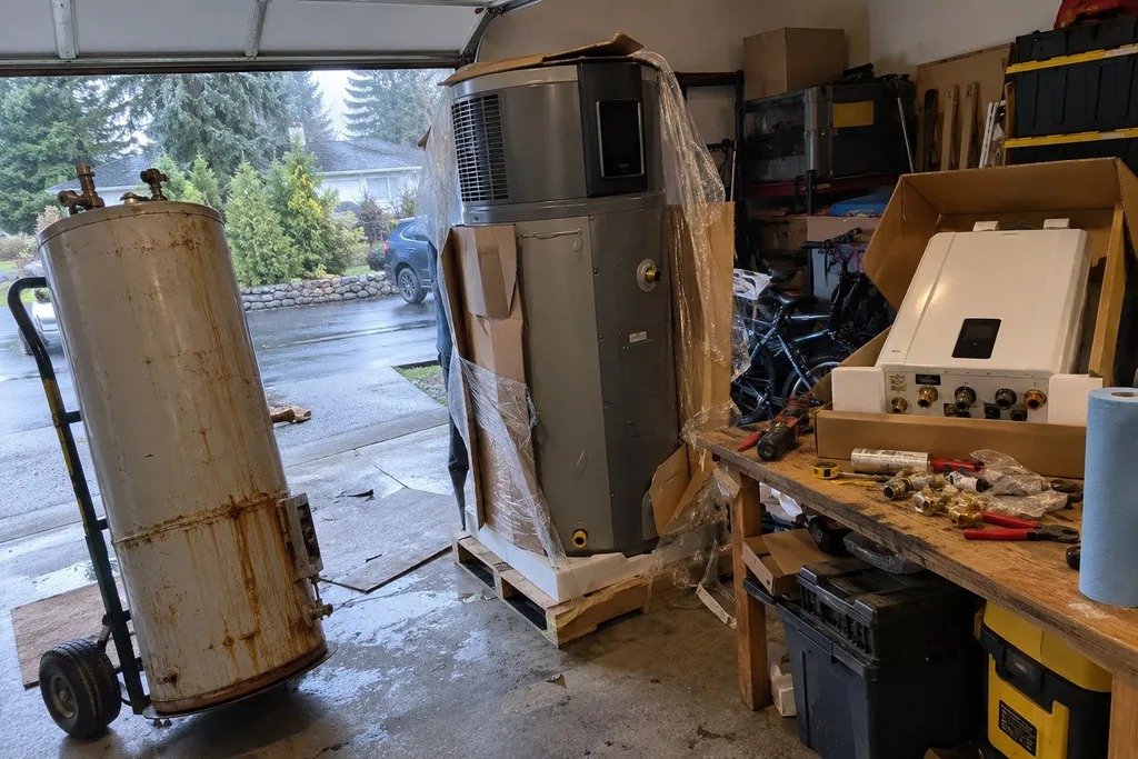 Old tank water heater on a hand truck beside a new heat pump unit and a boxed tankless heater in a Troutdale garage